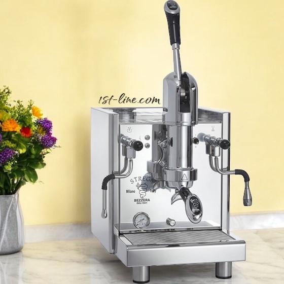 A Bezzera Strega S Tank V2 Espresso Machine with a polished stainless steel finish and dual steam wands sits on a countertop, with a vase of colorful flowers in the background against a pale yellow wall. © 2025 1st-line.com