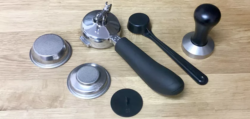 Accessories for the Rancilio Silvia M Espresso Machine displayed on a wooden surface, including a black-handled portafilter, metal filter baskets, a black measuring spoon, tamper, and a round black disk. © 2025 1st-line.com