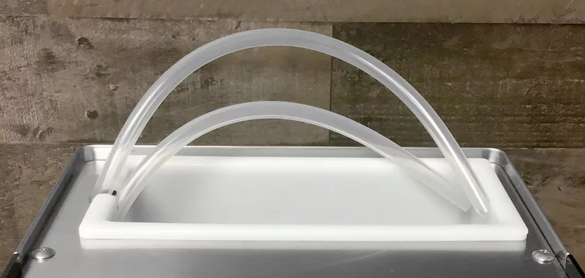 Two clear, flexible tubes rise from a white rectangular base placed next to a Rancilio Silvia M Espresso Machine on a metal counter with a brown tiled wall in the background. © 2025 1st-line.com