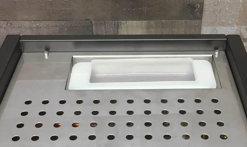 Close-up of a metal countertop with evenly spaced holes and an ice scoop holder, perfect for pairing with the Torre Peppina EVO Premium V1 Dual Boiler Espresso Machine w/ FC, set against wooden and stone panels. © 2025 1st-line.com